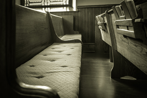 An empty wooden church pew with bibles. Photography in black and white with a horizontal orientation.