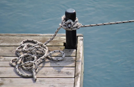A close up depiction of mooring, black and white rope attached to a black metal bollard on a wooden pier.