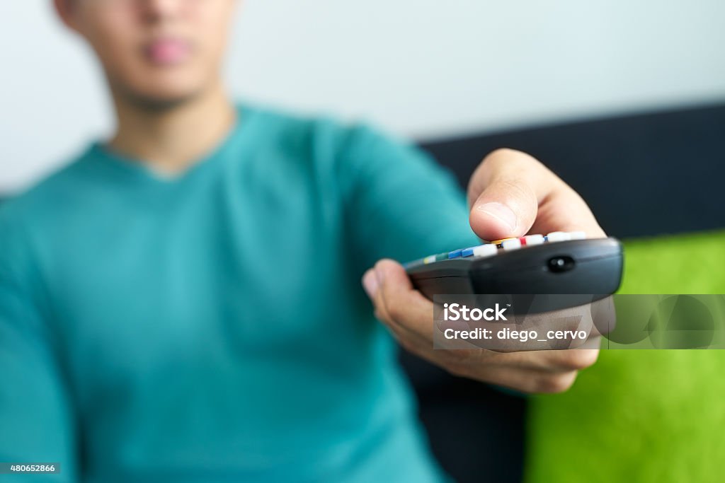 Asian Man Watches TV Changes Channel Holding Remote Control Asian young adult man watching TV and changing channel with remote control. Narrow focus on buttons and hand in foreground, cropped view. 2015 Stock Photo