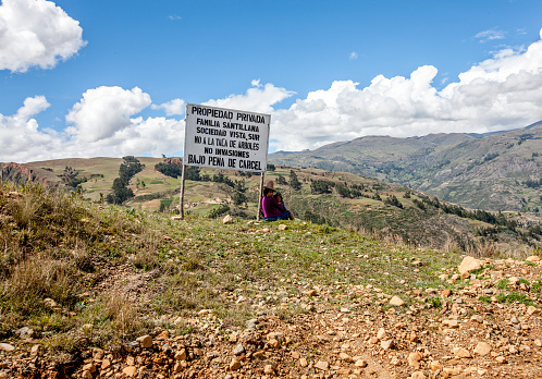 Huaraz, Peru - January 24, 2015: Campesina mother and her child in high Andes sitting under a sign while looking after her donkeys grazing on the hillside