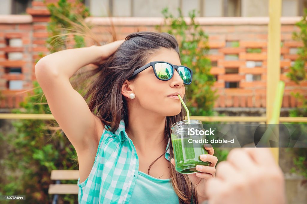 Woman with sunglasses drinking green vegetable smoothie outdoors Beautiful young woman with sunglasses drinking green vegetable smoothie with straw in a summer day outdoors. Healthy organic drinks concept. 2015 Stock Photo