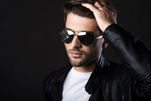 Handsome young man holding hand in hair and looking at camera while standing against black background