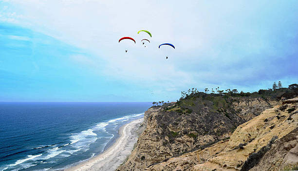 Paragliders over Black’s Beach in Early Summer Blacks Beach, San Diego’s famous clothing optional, nude beach, as seen from above the cliffs in early summer. Red, blue, and green paragliders float high in the sky while surfers, swimmers and sunbathers enjoy the afternoon surf and sun. Additional gliders can be seen near the distant horizon.  Image captured from Scripps Coastal Reserve. Curves have been adjusted to enrich and bring out details from the ocean and sky. torrey pines state reserve stock pictures, royalty-free photos & images