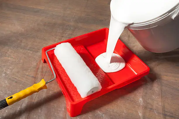 Pouring a white latex paint in a paint tray. A new paint roller is ready for use. A covered by plastic sheet floor is prepared for painting. Usually the cover prevents from unwanted dust and spots of paint during the renovation.  