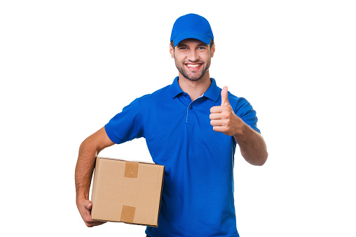 Cheerful young courier holding a cardboard box and showing his thumb up while standing against white background