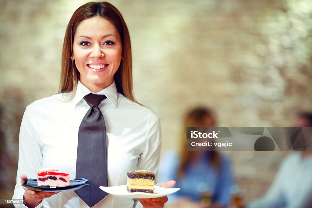 Cheerful waitress with desserts. Cheerful waitress with desserts looking at camera and smiling. Customers sitting in background. Waitress Stock Photo