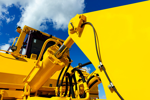 Bulldozer, huge yellow powerful construction machinery with big bucket, focused on hydraulic piston arm, blue sky and white clouds on background 