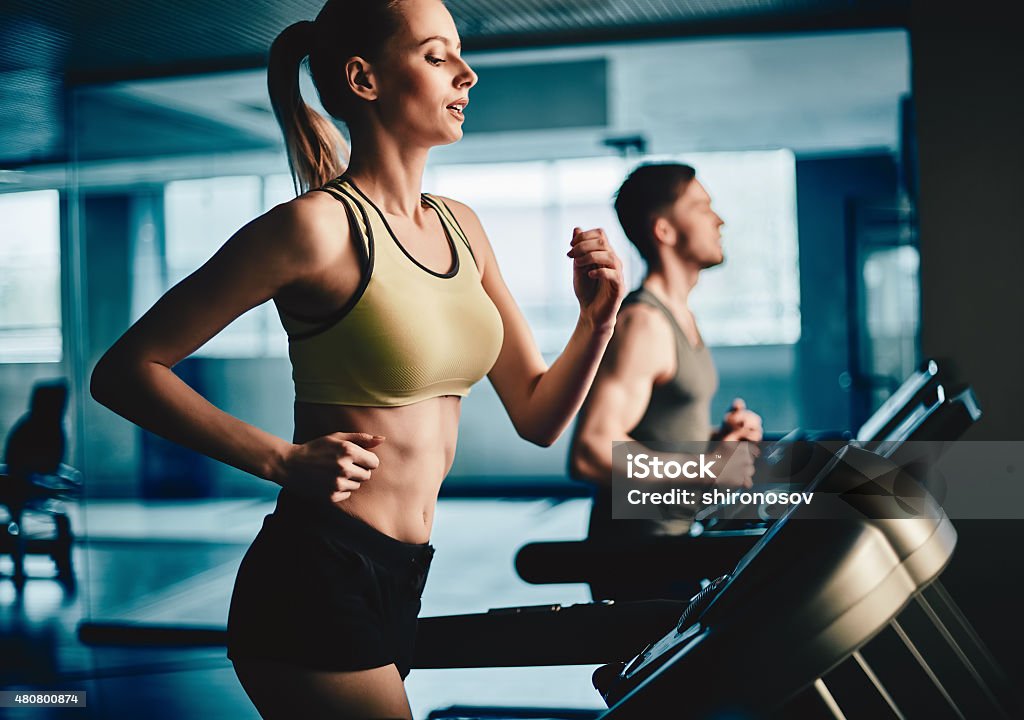 Woman on treadmill Active young woman running on treadmill with guy on background 2015 Stock Photo