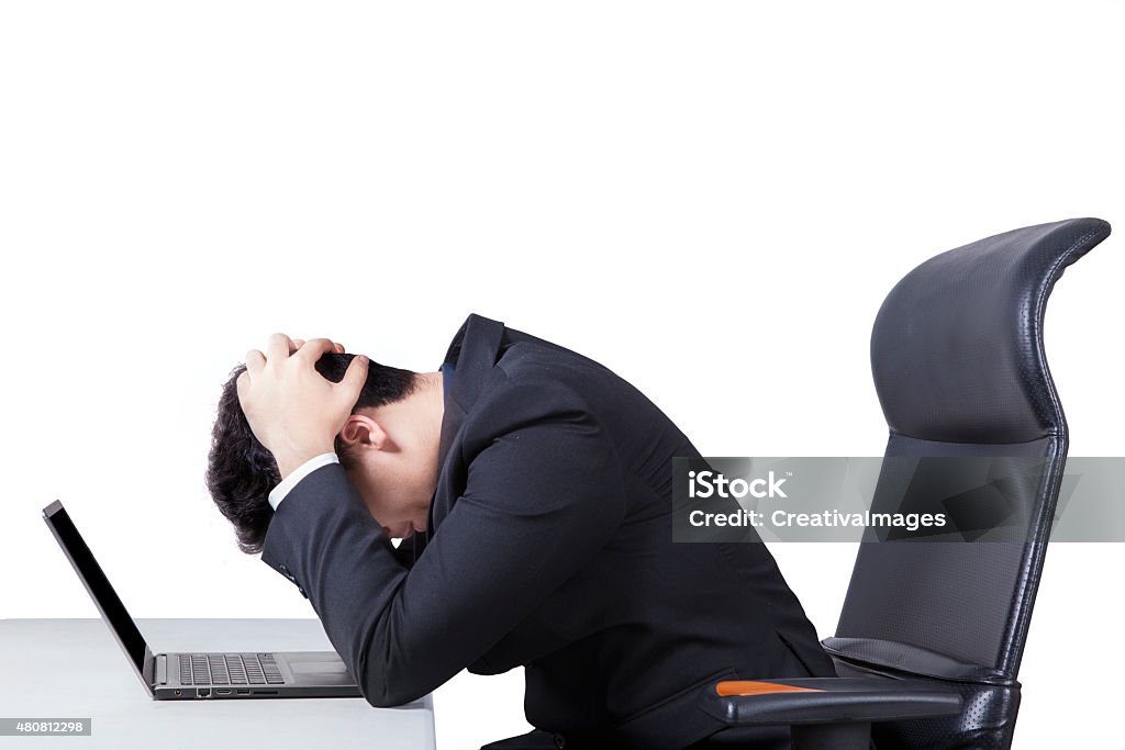 Confused worker with laptop on table Portrait of stressful male worker sitting on office chair while holding his head with a laptop on desk, isolated on white 2015 Stock Photo