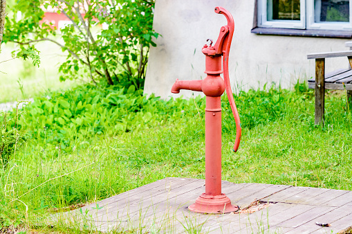 Red old iron water well pump with handle. Pump is placed on wooden lid outside building with green grass around it.