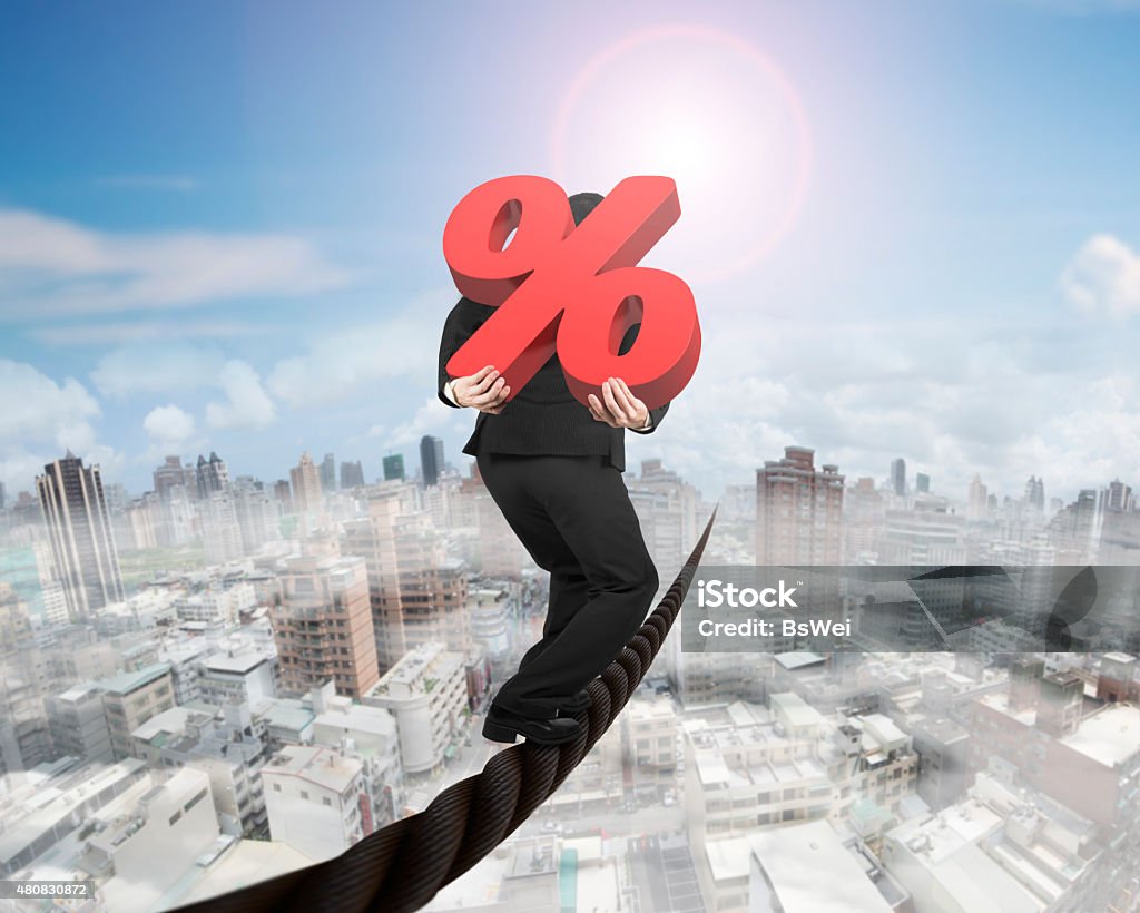 Businessman carrying 3D red percentage sign balancing on wire Businessman carrying 3D red percentage sign balancing on a wire, with sky sun mist cityscape background. 2015 Stock Photo