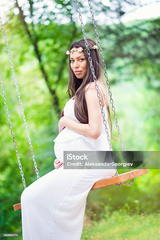 Beautiful Pregnant Woman Relaxing on Swing in Nature Beautiful brunette pregnant woman in white summer dress, sitting and relaxing on the swing in green natural park. 2015 Stock Photo