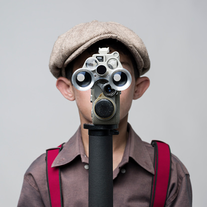 Little boy in old fashioned style filming via antique camera in front of gray background.The model is wearing flat hat.The camera in a position that it covers his face.The lenses of camera give eyes look on face.The photo was shot in studio with a medium format DSLR camera Hasselblad H4D in color.