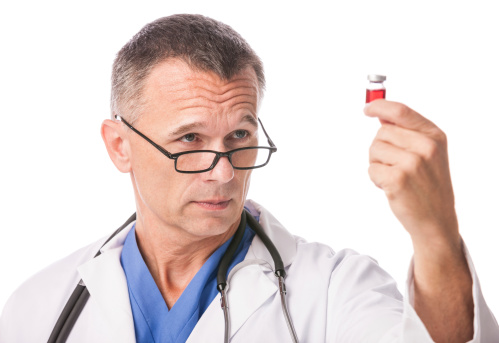 A mature caucasian male doctor or scientist holding up and examining a vial containing a red liquid. He is wearing a white lab coat and is isolated on a pure white background.