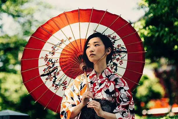 Beautiful japanese woman stand in the park. She wears kimono, obi and hold oil paper umbrella.The kimono is very colorful and elegant. She seems very pensive. 