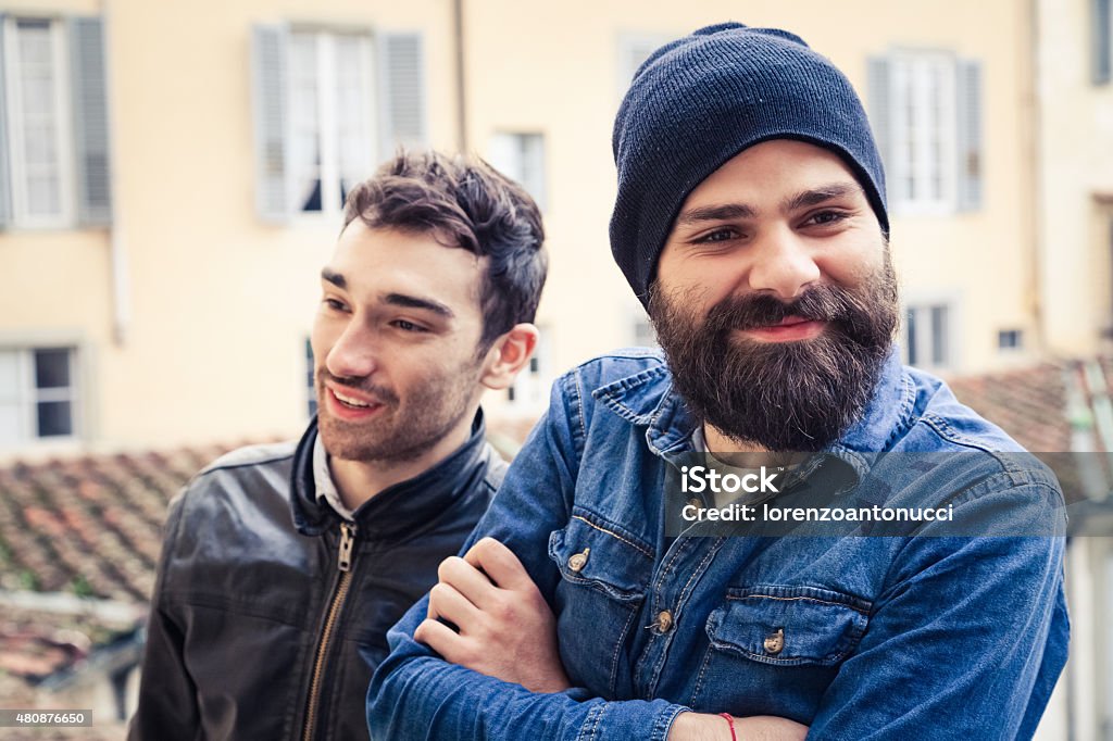 Couple of young men resting on the terrace A pair of two young men take a break from work in the terrace of a building. One has a beard, hat and a shirt, the other a leather jacket Beard Stock Photo