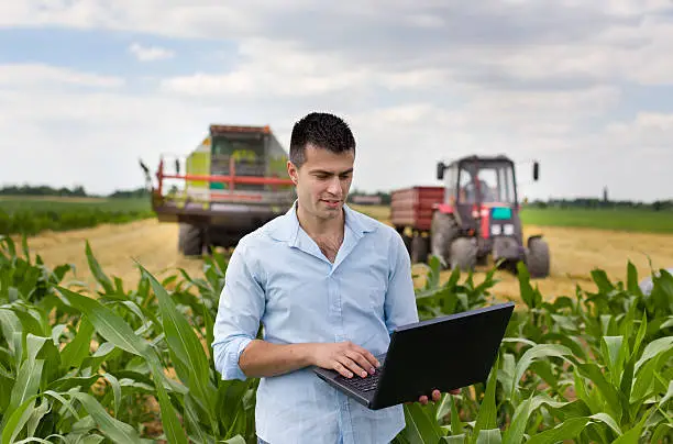 Young attractive farmer with laptop standing in corn field, tractor and combine harvester working in wheat field in background