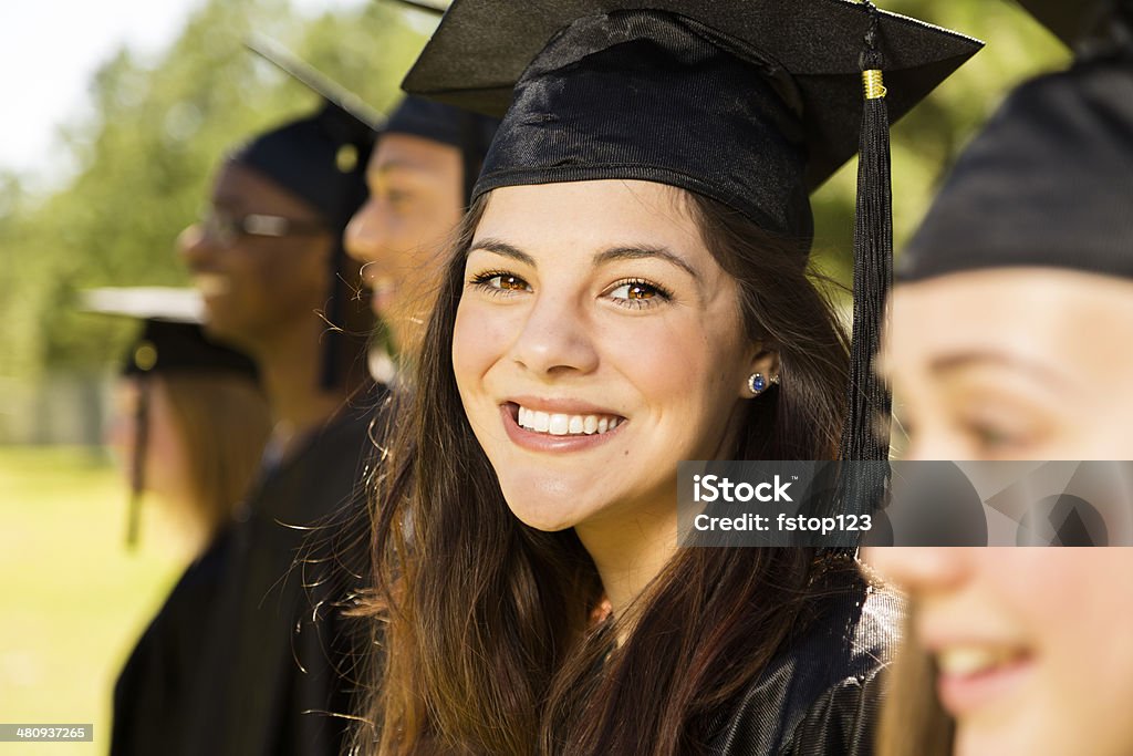 Education: Latin girl and friends at graduation. Cap, gown. Five, multi-ethnic friends dressed in cap and gowns excitedly wait for their name to be called during graduation ceremony during high school or college graduation. Latin girl looks at camera. Graduation Stock Photo