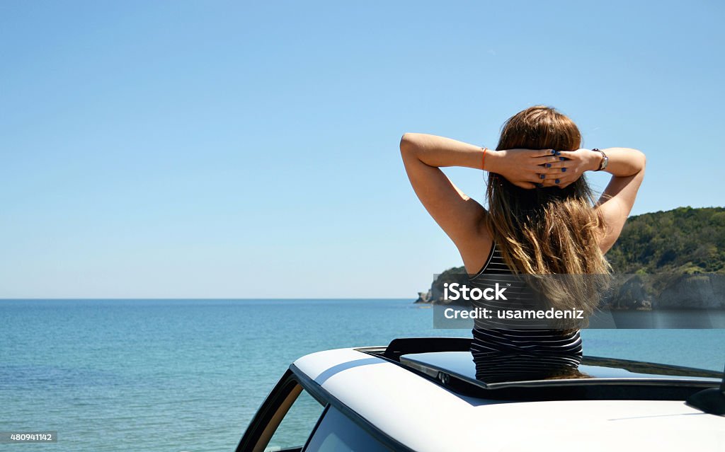 peace Rear view of relaxed woman on summer travel vacation to the coast leaning out car sunroof towards the sea. Sun Roof Stock Photo
