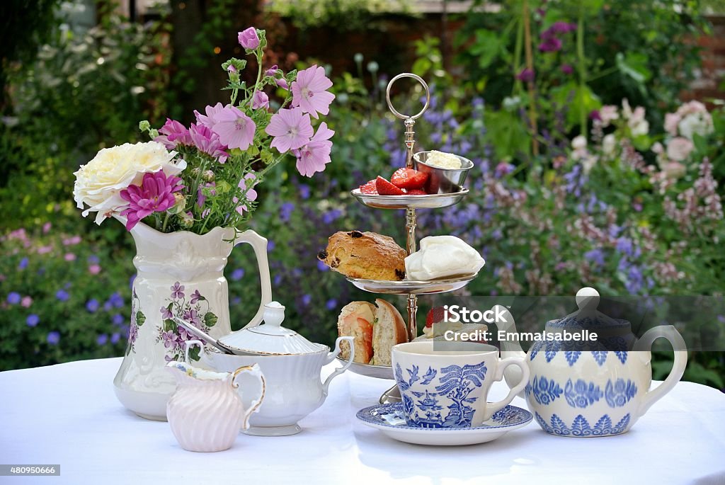 Afternoon Tea in a Country Garden Photograph of a table set for afternoon tea. The table is covered with a white table cloth and there is a three-tiered cake stand containing a sandwich, a scone, a meringue, a cake and some strawberries and cream. There is a blue and white teapot and cup and saucer and a white china milk jug and sugar bowl. To the left there is a jug containing an arrangement of flowers. The table is standing in an English country garden. Flowers can be seen in the background. Afternoon Tea Stock Photo
