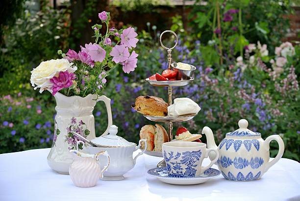 Afternoon Tea in a Country Garden Photograph of a table set for afternoon tea. The table is covered with a white table cloth and there is a three-tiered cake stand containing a sandwich, a scone, a meringue, a cake and some strawberries and cream. There is a blue and white teapot and cup and saucer and a white china milk jug and sugar bowl. To the left there is a jug containing an arrangement of flowers. The table is standing in an English country garden. Flowers can be seen in the background. garden parties stock pictures, royalty-free photos & images