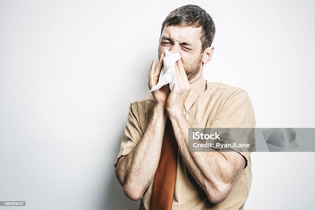 Man with Cold Sneezing A sick man with the common cold or allergies has a sneeze, a tissue covering his face.  Horizontal with copy space. Cold And Flu Stock Photo