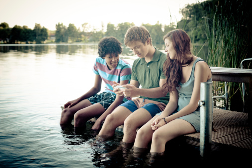 carefree summer day at lake: three young adults sitting on a jetty with their legs into the water