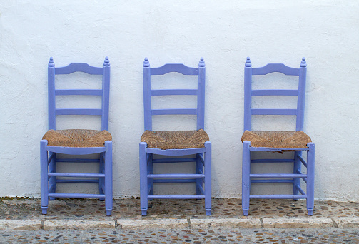 ¿where are our elderly people? 3 old blue wooden chairs alone in the mediterranean street on a white wall