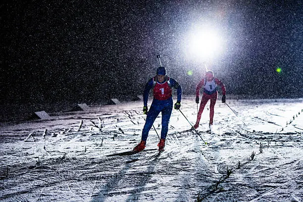 Male and female biathlete enjoying cross country skiing at night, they are carrying rifle on back.