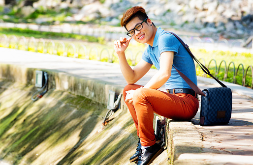 Young hip Vietnamese male student portrait in Hoan Kiem lake. He is sitting at the water's edge and lookin at the camera with a confident smile. He is wearing bright colored clothing and has red highlights in his hair. Photographed in Hanoi, Vietnam.