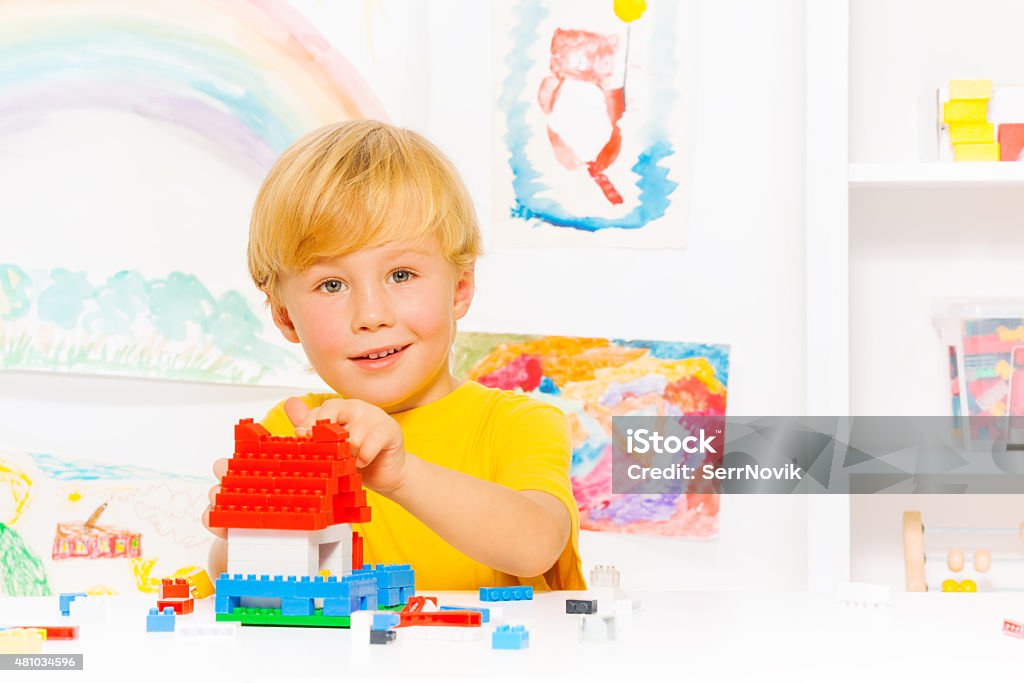 Handsome blond boy building house Close portrait of blond little preschool boy in glasses playing with plastic blocks constructing simple house in the room background 2015 Stock Photo