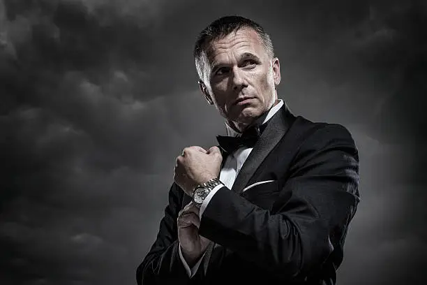 A handsome mature male secret agent, bodyguard, spy, or security staff dressed in an elegant tuxedo and bow tie as he adjusts his watch or cuff links on a stormy night with cloudy sky in the background.
