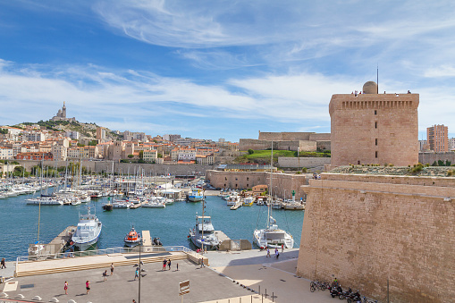 Marseille, France - August 24, 2014: Panoramic view of Marseille from Fortress  - Basilique Notre-Dame de la Garde , Fort Saint Jean and Vieux Port of Marseille. Many people walking on promenade. Many ships at pier. In background the cityscape of Marseille and Basilique of Notre Dame de la Garde. On the right some people on tower and fortress.