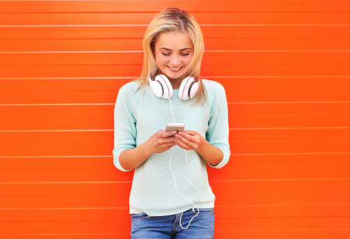 Fashion, technology and people concept - pretty smiling girl using smartphone in headphones against the colorful orange wall