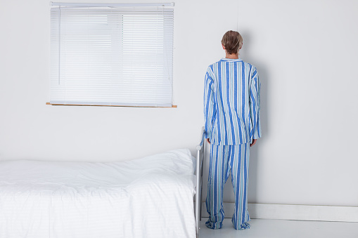 A tall, slender man in his 30s wearing traditional, striped pyjamas about to go to bed in his stark and monastic bedroom. He is standing with his back to the camera and it's unclear whether he is emotionally normal.