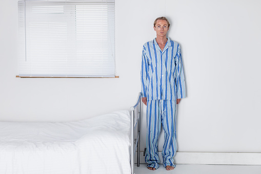 A tall, slender man in his 30s wearing traditional, striped pyjamas about to go to bed in his stark and monastic bedroom. Post-processed for drama.