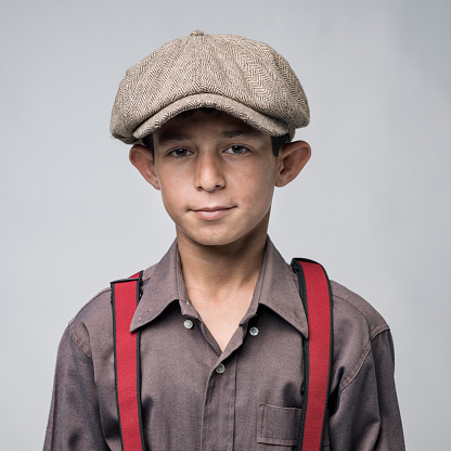 Head and shoulders portrait of little boy wearing a brown shirt, red suspenders and a flat newsboy cap in front of gray background.The model has dark hair and smiling.The photo was shot with a medium format camera Hasselblad H4D in studio.