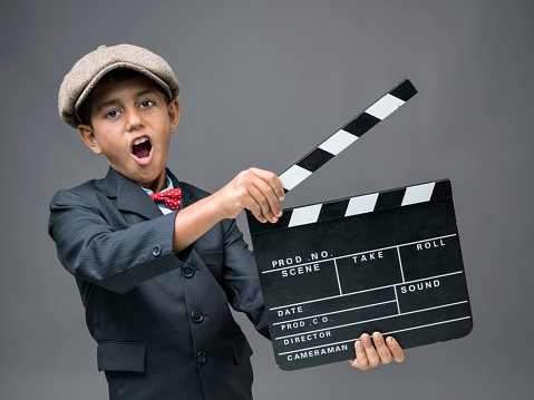 Mixed race little boy wearing a black suit a brown flat cap and red bow tie holding film slate and shouting in front of dark gray background.The model is standing on the left side of frame and his mouth is wide open.The image was shot with medium format camera Hasselblad H4D in studio.