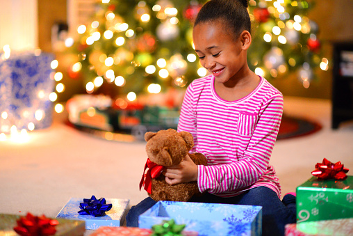 A little girl opening a teddy beard on Christmas morning in the family living room next to the Christmas tree.