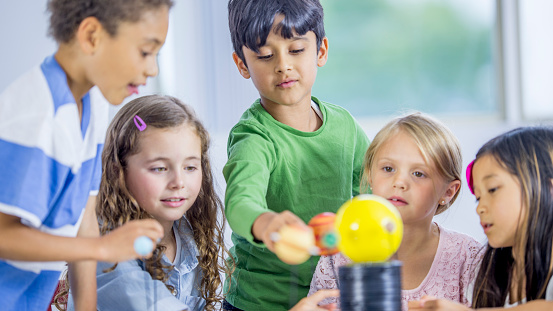 A multi ethnic group of little children sitting at a desk, exploring a model solar system for a science class.