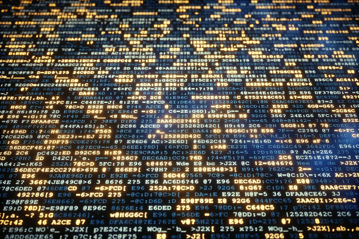 An abstract IT background concept representing encrypted data on a flat shiny surface. Random blocks of data are organized in rows and come in a blue and yellow hue. Some characters are glowing more brightly.