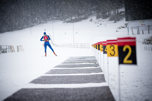 Male biathlete wearing skiwear and standing on snow, rifle on back.