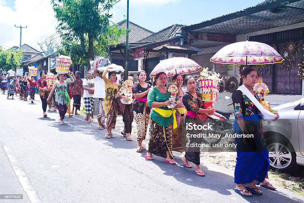 Balinese people with donations walking along the road. Bali, Indonesia - June 21, 2015: Part of traditional Balinese cremation ceremony. Balinese people, dressed in traditional clothes walking by the road with donations. Sunny day in the village near Ubud. 2015 Stock Photo