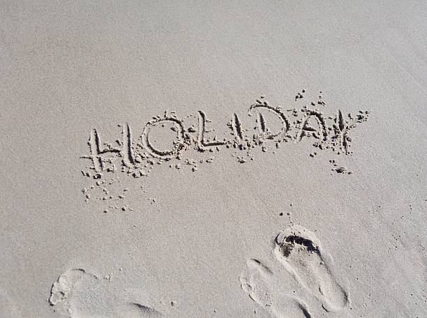 Holidays is the key word written in the golden sand with blue sea behind