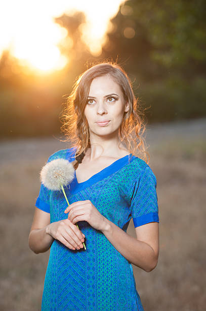 Portrait of brunette young woman in blue dress holding a big dandelion in warm sunset. Summer, outdoors. Beautiful girl with dandelion enjoying the summer sun outdoors in the park