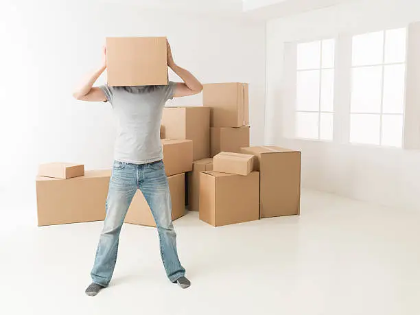 man hiding his head in cardboard box, standing in new apartment, despair gesture