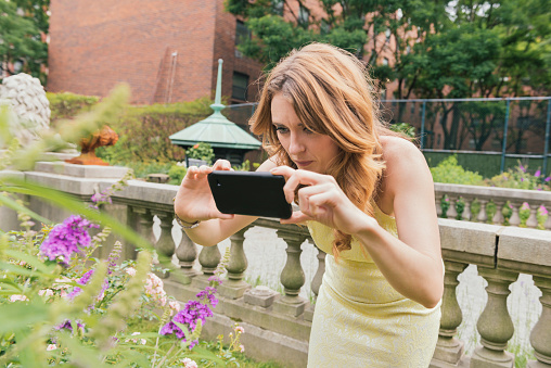 This is a horizontal, color, royalty free stock photograph of a happy, white smiling British feminine woman living in New York City. She spends time in urban Manhattan on a summer day in the SoHo area. In a yellow dress she stops to photograph the colorful flowers with her mobile phone. Photographed with a Nikon D800 DSLR camera.