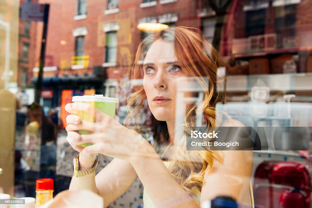 NYC Woman Drinking Coffee Looks Out Window at Cafe Restaurant This is a horizontal, color, royalty free stock photograph of a white British feminine woman living in New York City. She spends time in urban Manhattan on a summer day in the SoHo area. She sits at a small cafe restaurant. She sits at a counter holding a cup of coffee and looking out a window facing the sidewalk. The architecture from the buildings across the street are reflected on the window. Photographed from outside with a Nikon D800 DSLR camera. New York City Stock Photo
