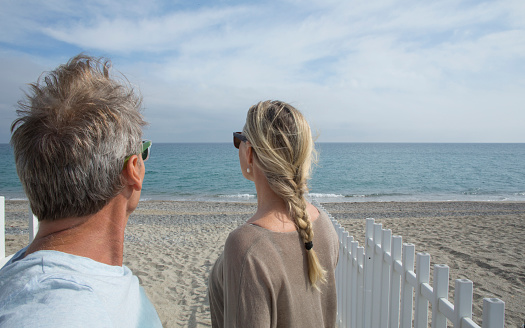 Couple look out at the Mediterranean in Liguria, Italy. With white fence and beach in foreground.