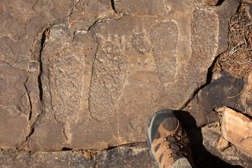 Petroglyphs of two pairs of feet stand (and another petroglyph pair out of camera) on top of a large boulder and face towards an Apishapa Native American fortified village known as the Sorensen site (1276 - 1299 A.D.), 700 to 800 years old which stands over the Picket Wire Canyonlands along the Purgatoire River in the Comanche National Grasslands of southeast Colorado.